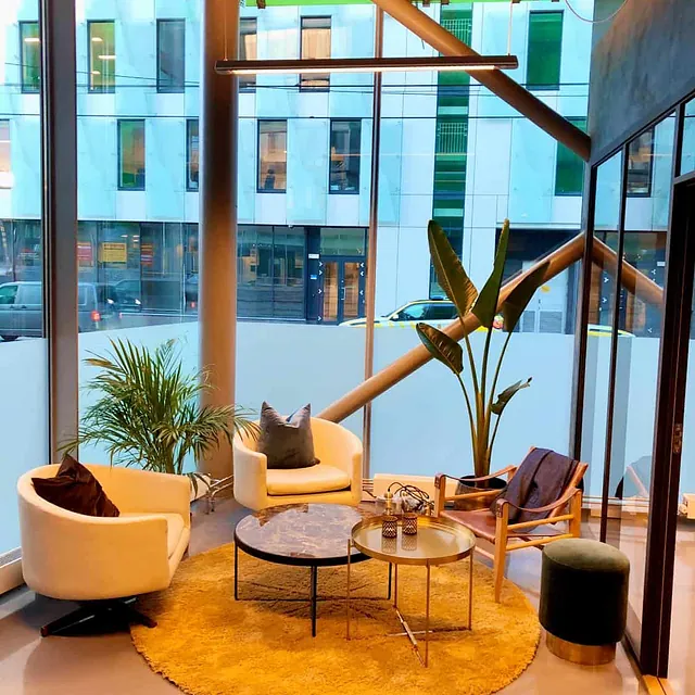 Moderne loungeområde med lenestoler, planter og runde bord ved store vinduer.