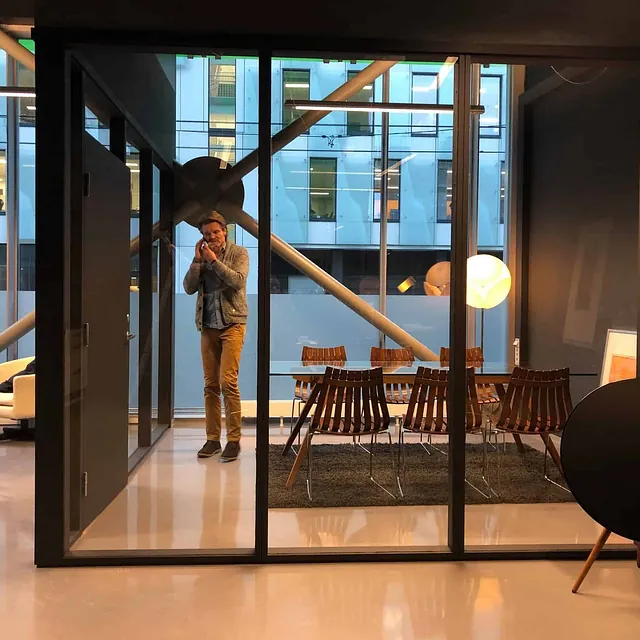 Mann i telefon i glassinnrammet møterom med designstoler og lamper.