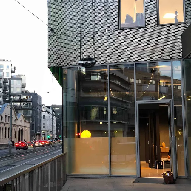 Inngangsparti til kontorbygg med glassfasade og utsikt mot gate med trafikk.
